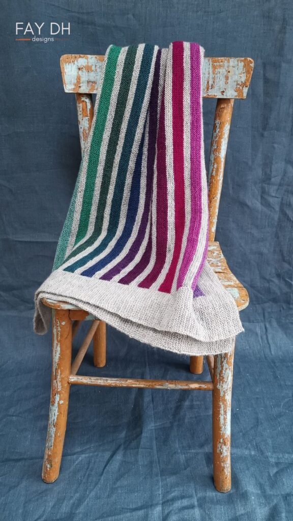 Striped Scottish Rainbow blanket on a chair. In muted heather and sea tones with a light grey main colour.