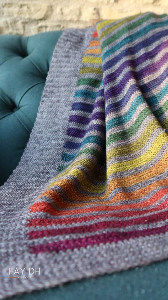 Striped Stormy Rainbow blanket on a chair. In earthy rainbow tones with a dark grey main colour.