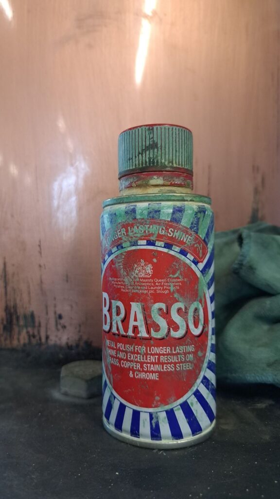 Bottle of Brasso with red, white and blue and a copper still in the background with a cloth in verdigris.