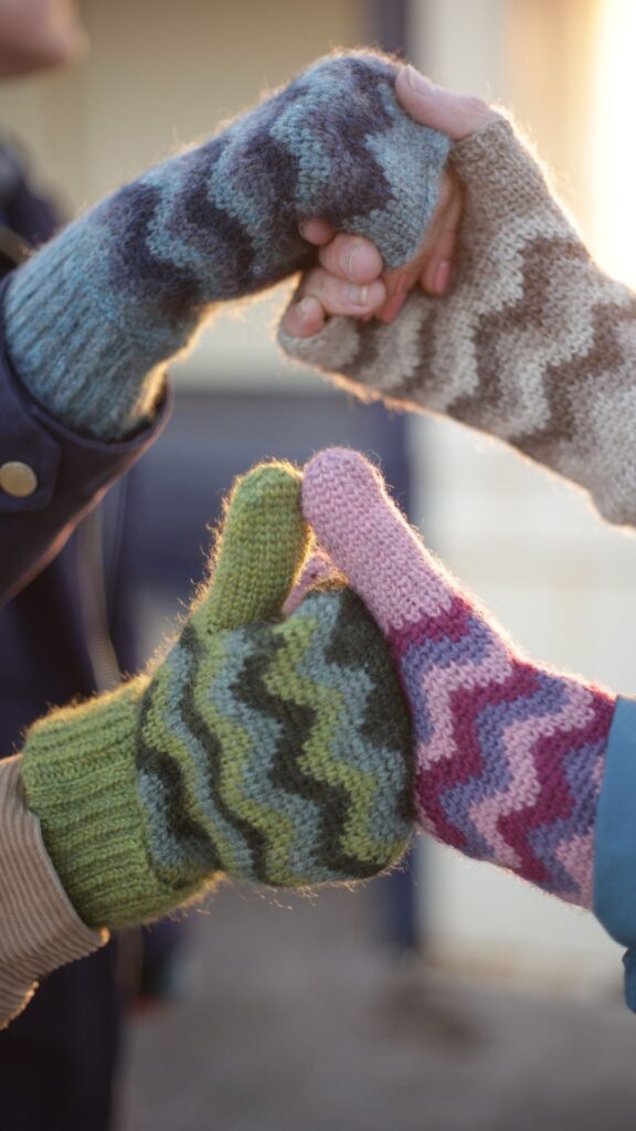 Hands wearing crocheted mittens. One green pair and one pink and purple, one in blues and another in natural wool colours. All with crocheted colourwork wave patterns.