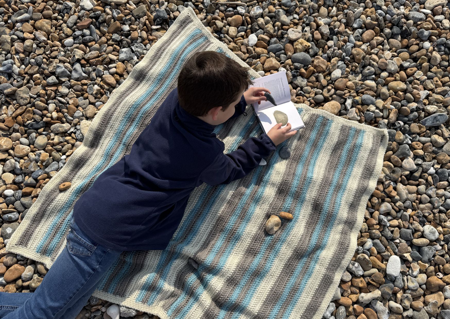 Striped crocheted Pebble Spotter blanket on a pebbled beach with a small boy on it identifying pebbles.