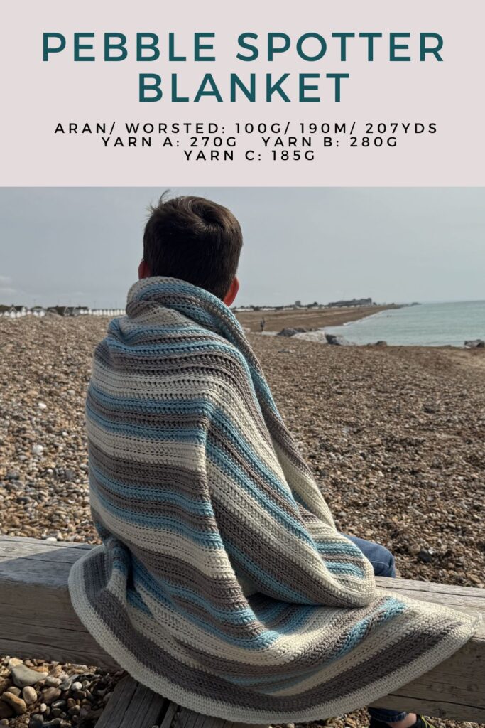 Striped crocheted Pebble Spotter blanket wrapped around a small boy on a pebbled beach.