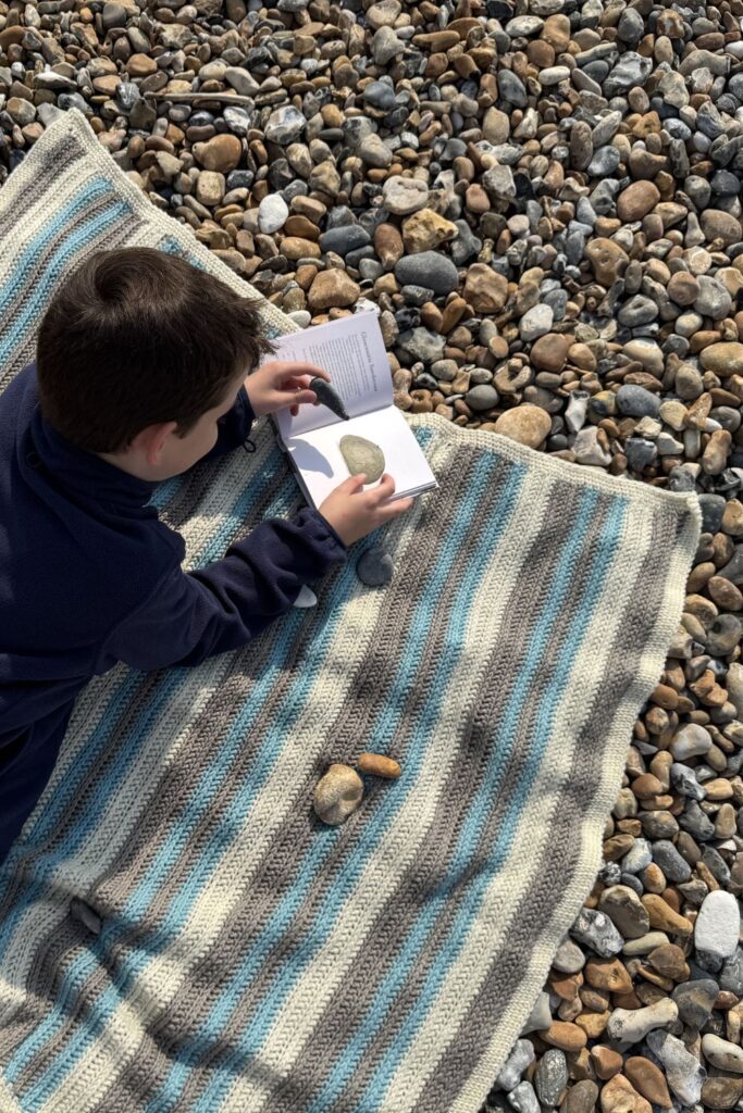 Striped crocheted Pebble Spotter blanket on a pebbled beach with a small boy on it identifying pebbles.
