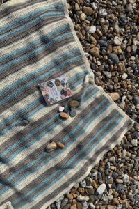 Striped crocheted Pebble Spotter blanket on a pebbled beach.